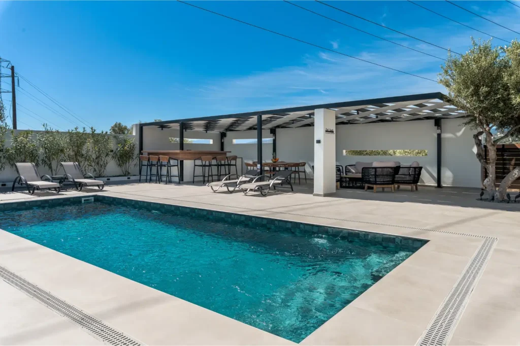 Luxury outdoor swimming pool with sun loungers, a pool bar, and a shaded lounge area under a clear blue sky at Ria's Apartment.