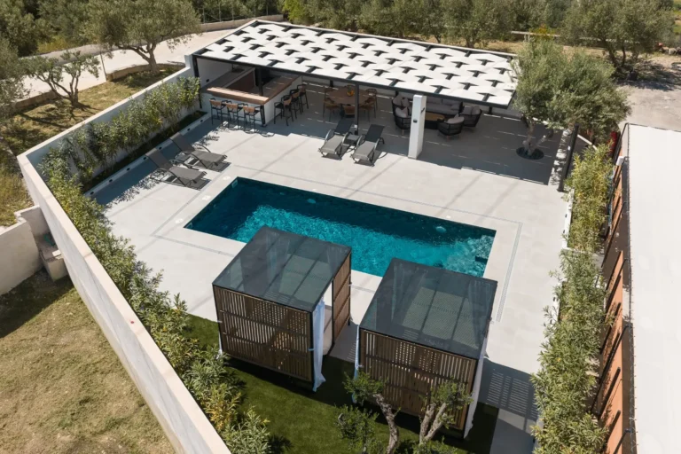 Aerial drone shot showing the entire outdoor area of Ria's Apartment, featuring the pool, cabanas, pool bar, and dining area under the pergola.
