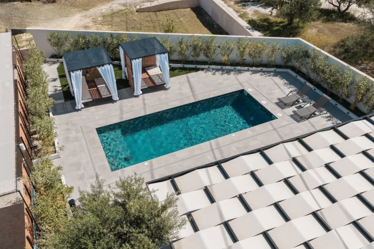 Panoramic drone shot showing the swimming pool, luxury cabanas with white curtains, and the shaded area of Ria's Apartment during the day.
