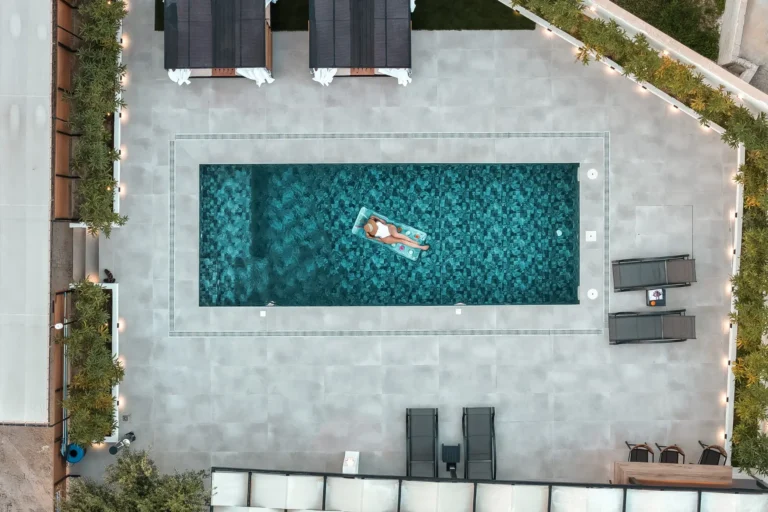 Overhead drone shot of the illuminated swimming pool at Ria's Apartment at night, featuring a woman on a pool float and the surrounding outdoor area.