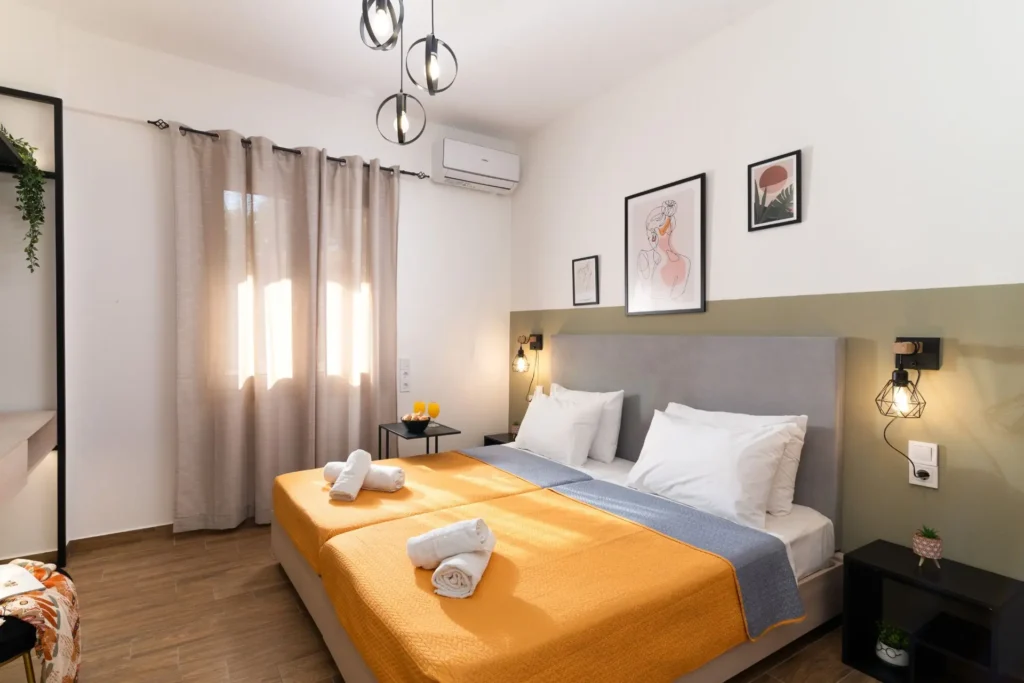 Comfortable double bed with yellow and grey covers, a grey tufted headboard, and geometric bedside lamps at Ria Oasis Residence in Agios Nikolaos.