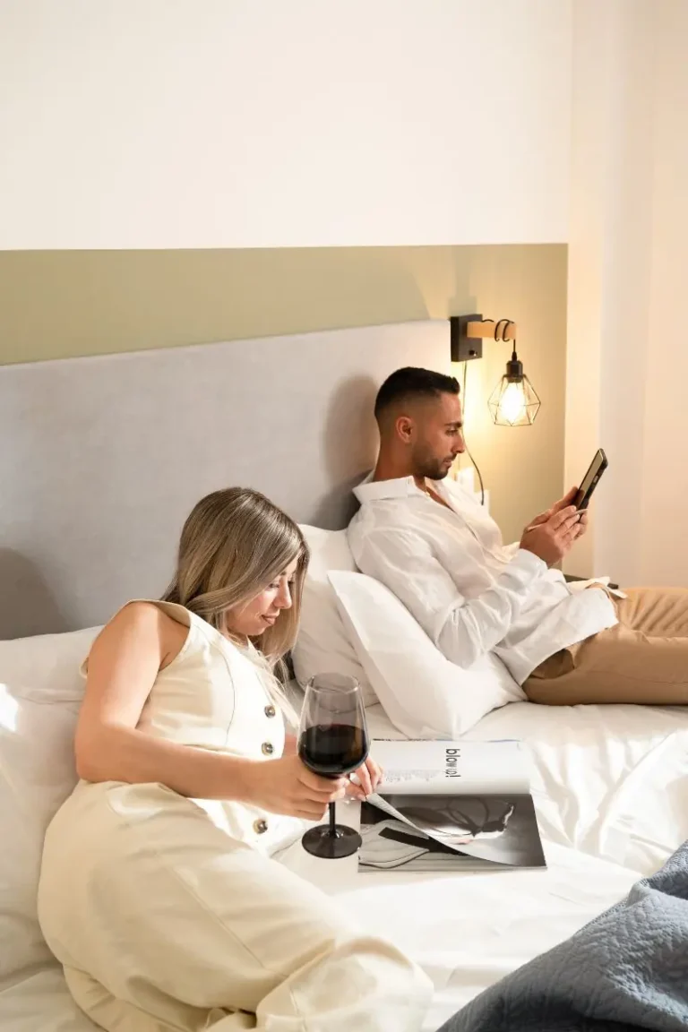 A couple relaxing in a modern bedroom at Ria's Apartment in Agios Nikolaos, reading a magazine and using a smartphone.