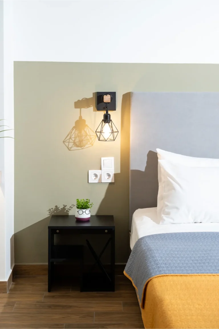 Close-up of a bed at Ria's Apartment featuring a grey headboard, industrial wall lamp, black nightstand, and wooden flooring.