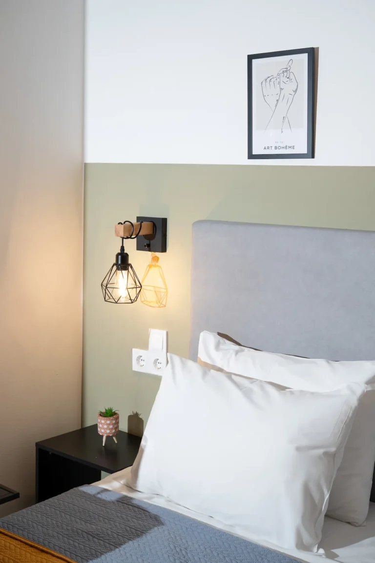 Bedroom detail at Ria's Apartment featuring an industrial wall lamp, an "Art Bohème" framed print, a grey headboard, and a small plant on a nightstand.