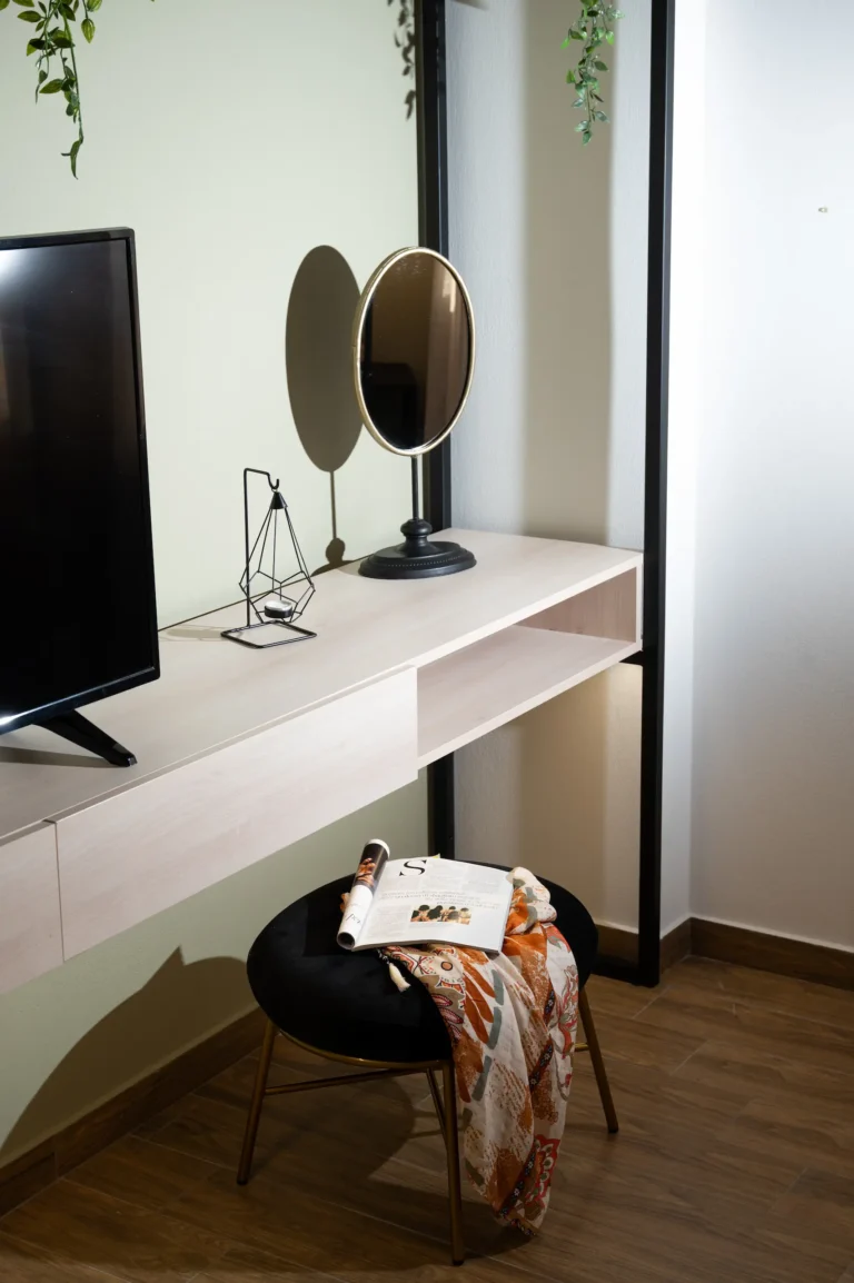 A modern wooden vanity desk in a bedroom, featuring an oval mirror, a black velvet stool, and decorative items.