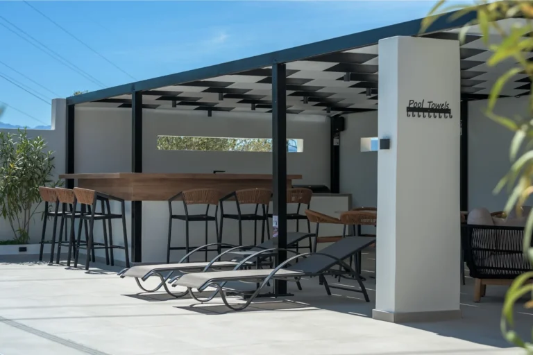 Elegant pool bar with a wooden counter and high stools under a modern pergola, with sun loungers in the foreground at Ria's Apartment.