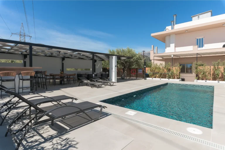 Panoramic view of the outdoor swimming pool, pool bar, and the building of Ria's Apartment under a clear blue sky.
