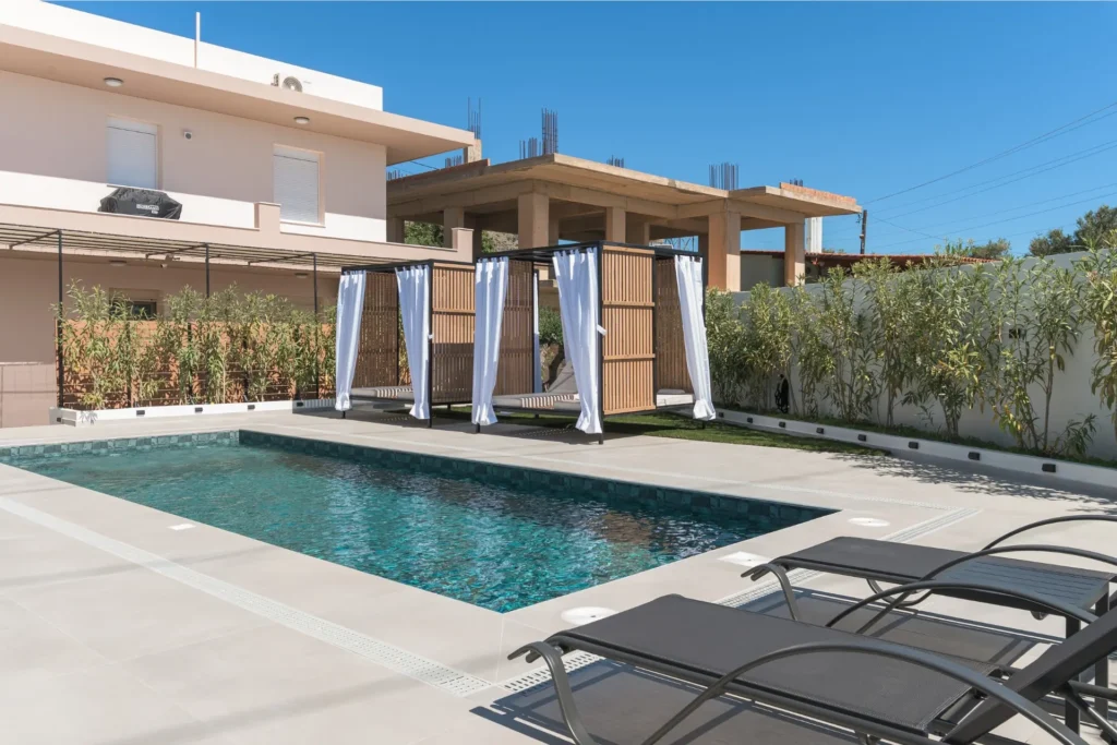 Outdoor swimming pool featuring modern sun loungers in the foreground and luxury wooden cabanas with white curtains at Ria's Apartment.