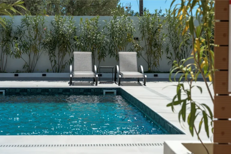 Two modern sun loungers with a small table between them, positioned at the edge of the swimming pool with a background wall of oleander plants at Ria's Apartment.