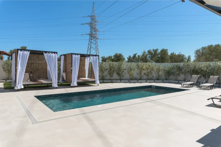 Modern outdoor swimming pool with luxury cabanas featuring white curtains and sun loungers under a clear blue sky at Ria's Apartment.
