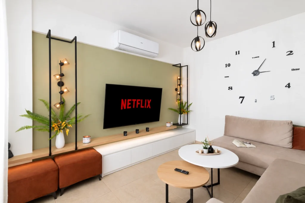 Spacious, modern living room at Ria's Apartment in Agios Nikolaos, featuring a large wall clock, comfortable sofa, and Netflix on TV.