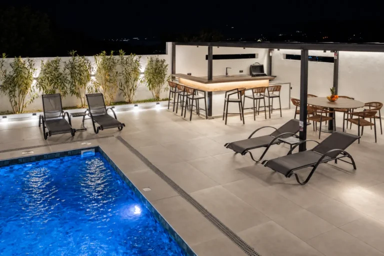 Illuminated outdoor swimming pool at night with a covered patio, bar, dining area, lounge seating, and sunbeds at Ria Oasis Residence.