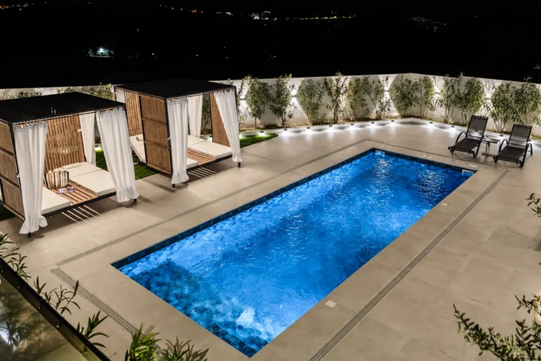 Illuminated outdoor swimming pool at night with luxury poolside cabanas, lounge chairs, and wall-mounted nighttime lighting at Ria Oasis Residence.