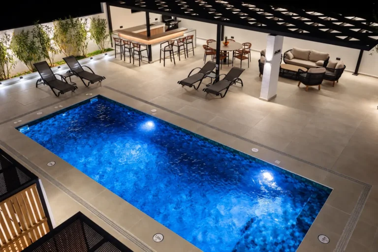 Night view of the illuminated swimming pool at Ria's Apartment, featuring the pool bar, dining area, and outdoor lounge highlighted by warm lighting.