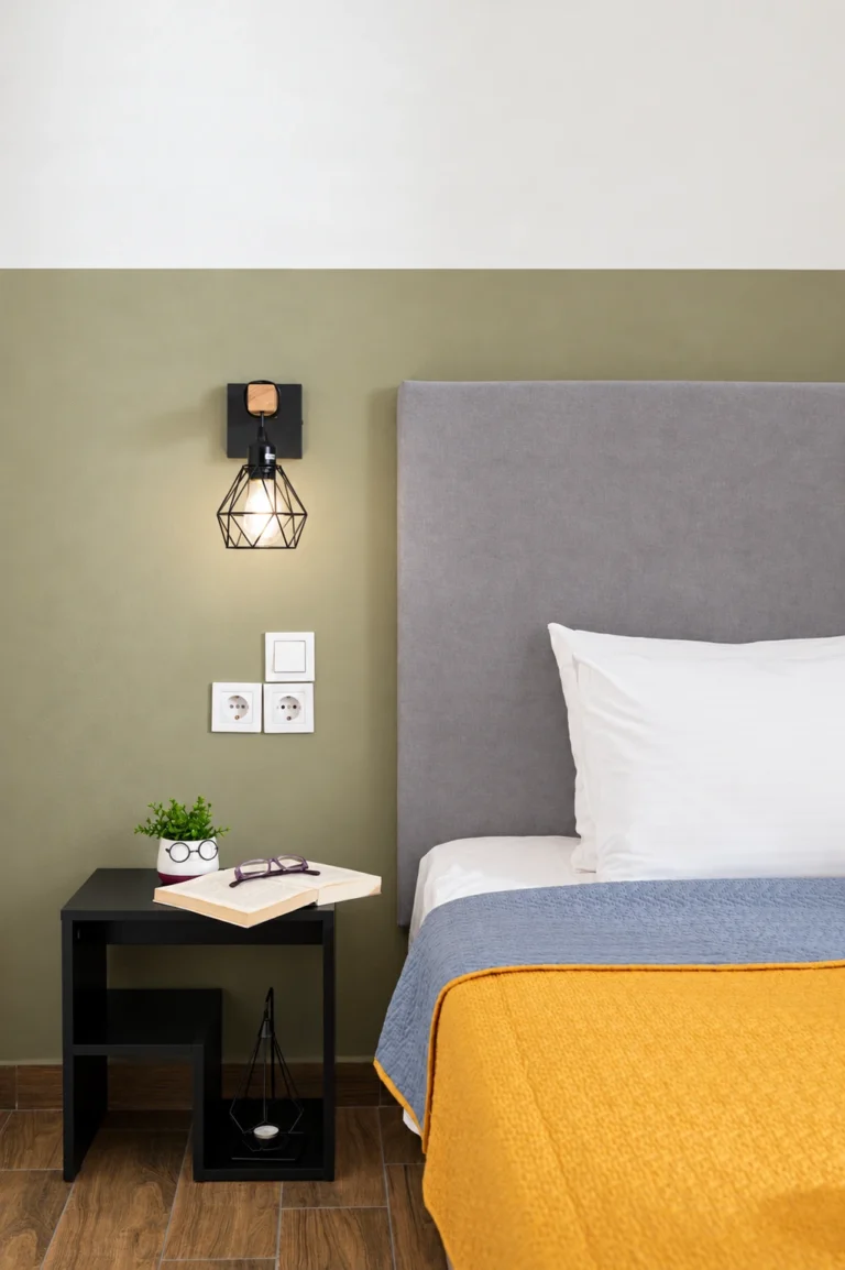 Close-up of a modern black nightstand with an open book and a geometric wall lamp next to a bed at Ria Oasis Residence in Agios Nikolaos.