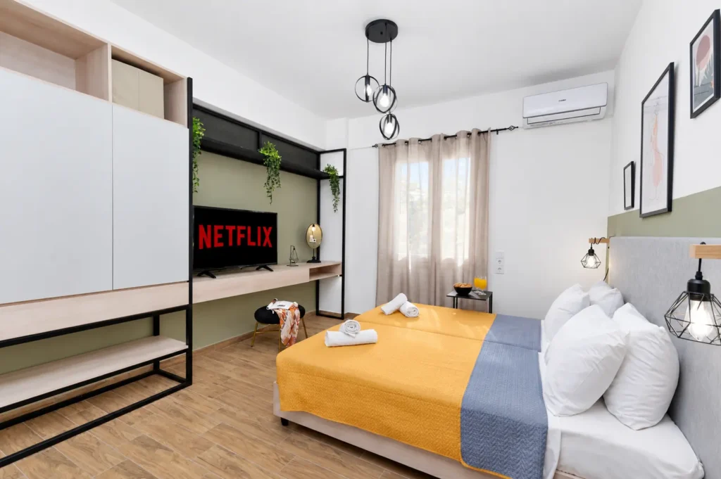 Bright master bedroom with a double bed, yellow covers, Netflix on TV, and modern wardrobe at Ria Oasis Residence in Agios Nikolaos.