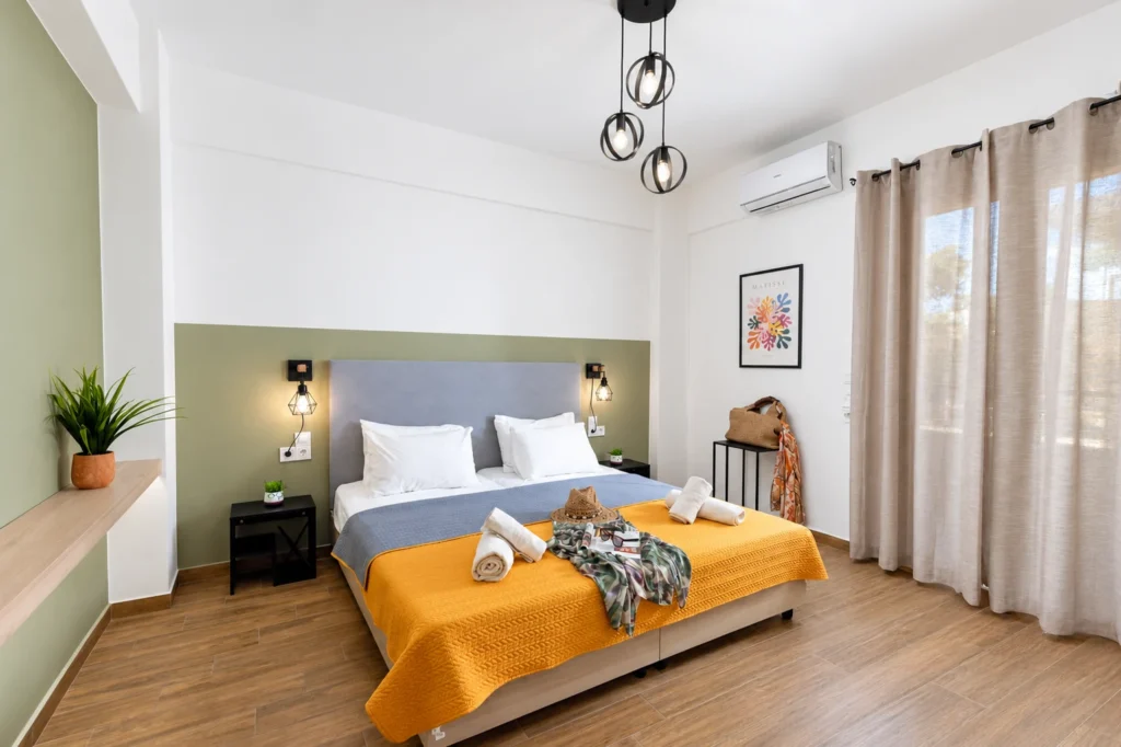 Modern second bedroom with a double bed, bright yellow and grey bedding, and a balcony door at Ria Oasis Residence in Agios Nikolaos.