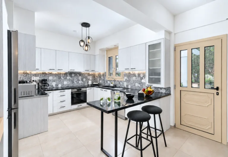 Φωτεινή κουζίνα του Ria Oasis Residence στον Άγιο Νικόλαο με μπαρ, δύο σκαμπό, πλυντήριο πιάτων, παράθυρο και πόρτα προς την αυλή